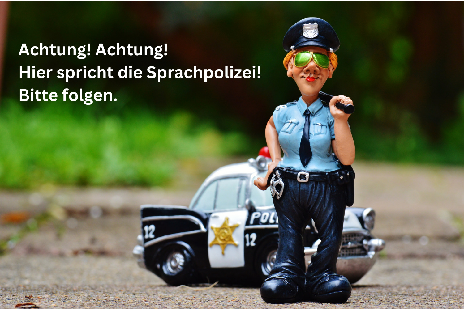 Polizeifigur in Uniform mit Sonnenbrille, weiblichen Rundungen und Mikro in der Hand. vor sehr kleinem Polizeiauto iun Landschaft, ironische Darstellung. Text: Achtung! Achtung! Hier spricht die Sprachpolizei! Bitte folgen.