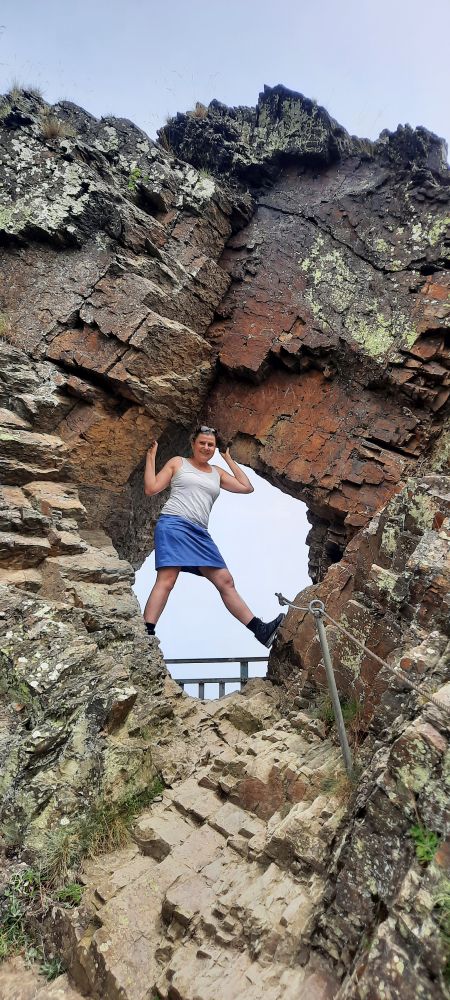 Felsen mit Loch. Darin steht Frau in sommerlicher Wanderkleidung und lacht in die Kamera. Sichtbar auch ein Metall-Seil, was beim Erklimmen des Felsens Halt gibt.