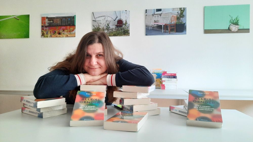 Frau mit langen braunen Haaren lächelt in die Kamara, das Kinn auf die Hände gestützt, die auf einem Buchstapel liegen. Buch "Alle(s) Gender. Wie kommt das Geschlecht in den Kopf?". - Im Hintergrund hängen Kunstfotografien an der Wand.